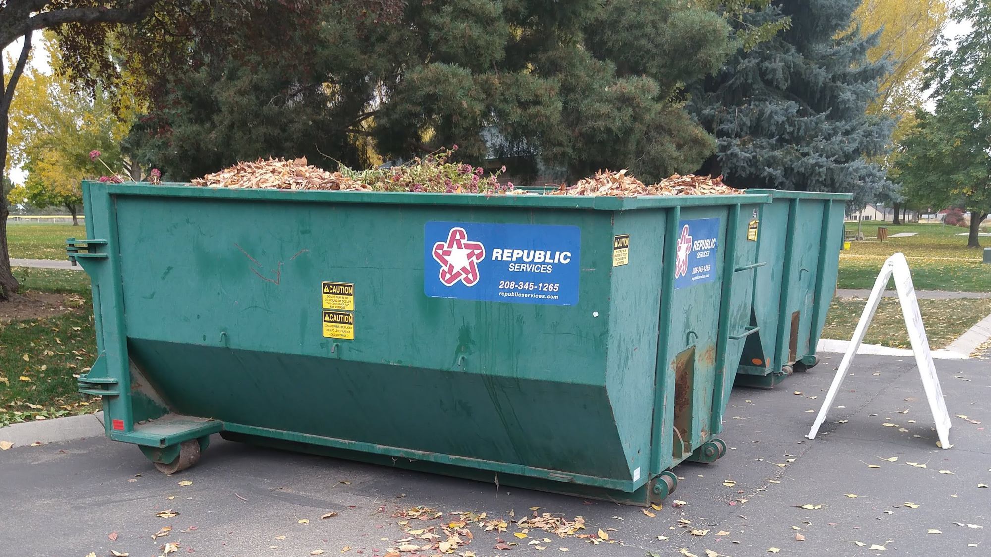 Two large, green metal bins full of leaves at a park with large trees in the background
